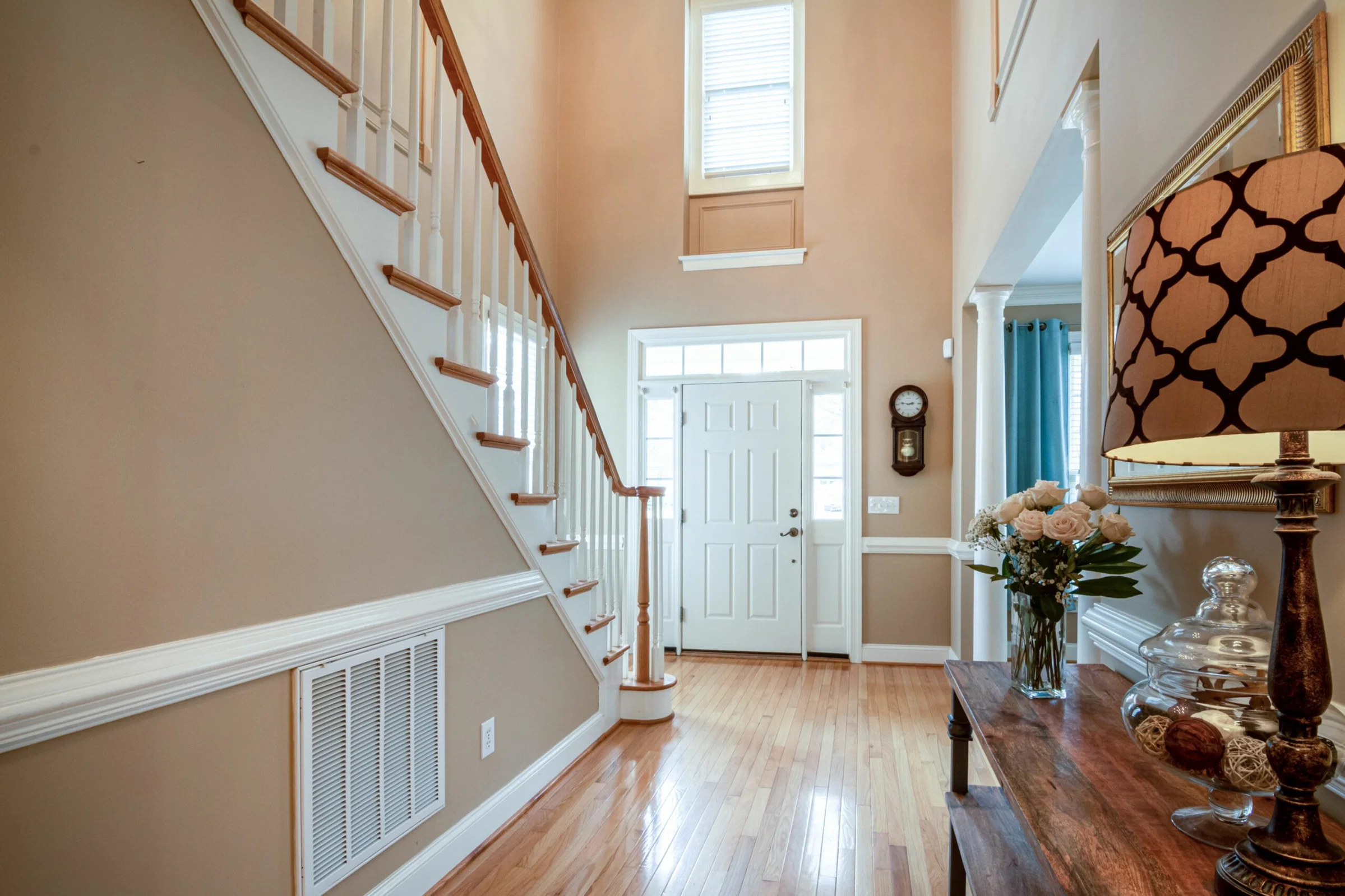 Bright foyer with a white front door, staircase on the left, and tall windows letting in daylight; floral arrangement on a wooden table nearby.