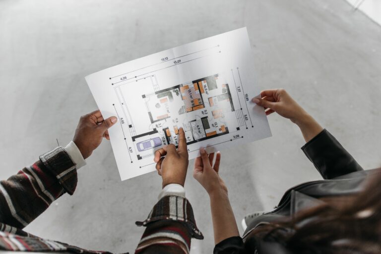 Two people hold and examine a detailed architectural floor plan outdoors, showing rooms and measurements.