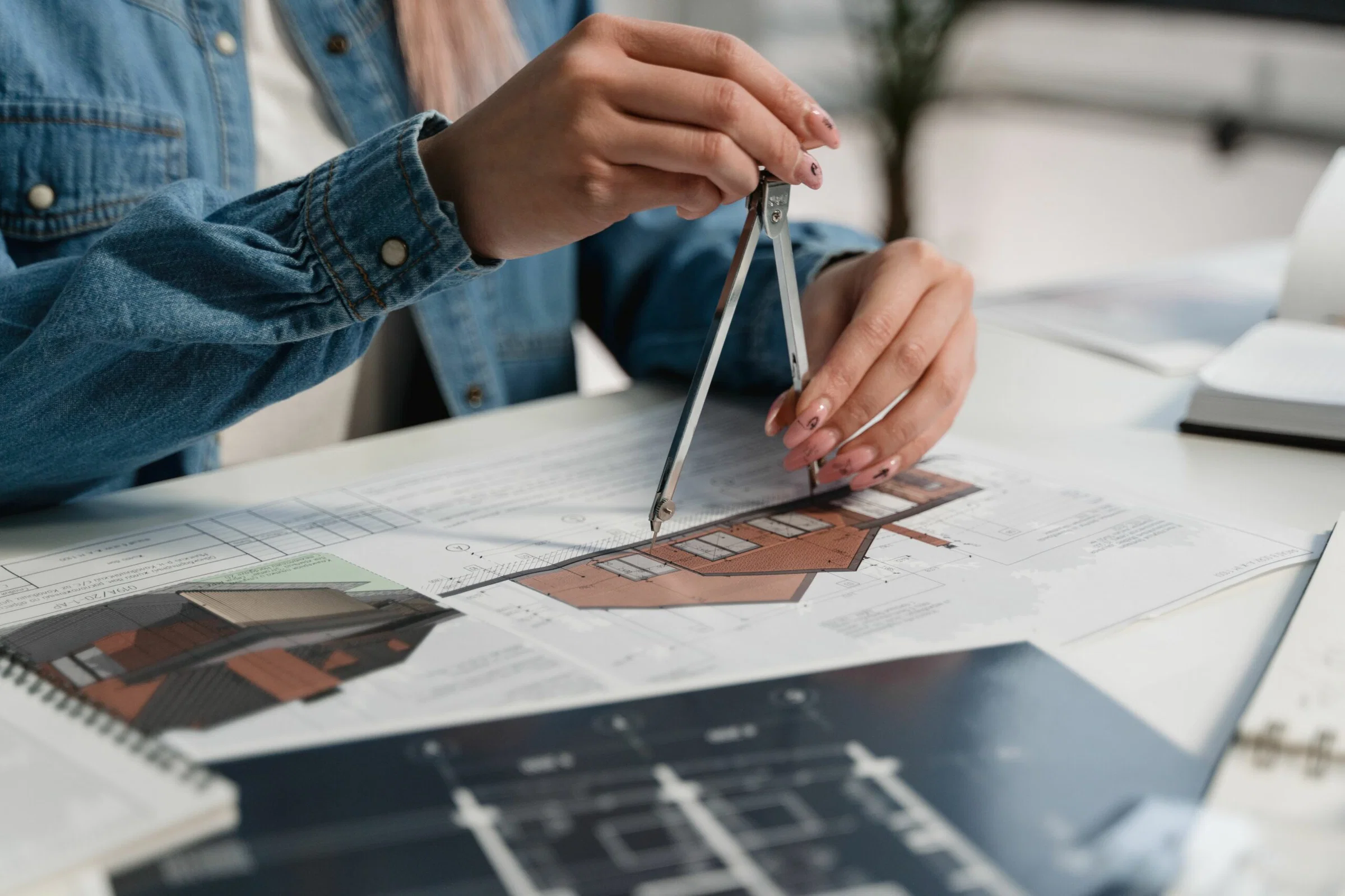 Person in a denim jacket uses calipers to measure a building plan on a desk, focusing on the hands and tools.