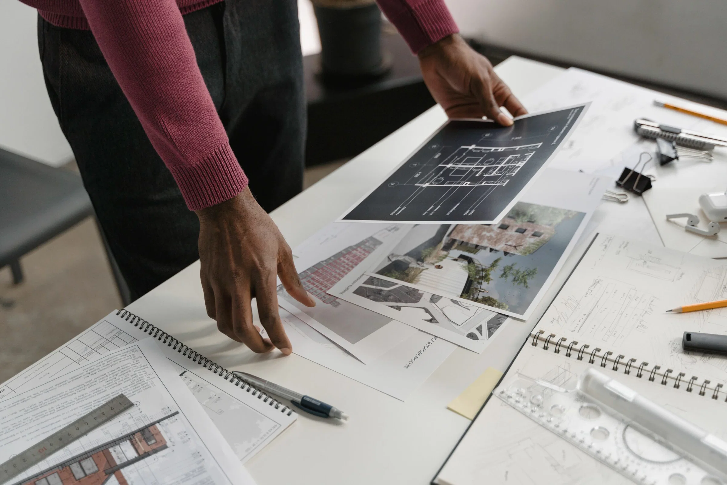 Person in a maroon sweater reviews architectural blueprints spread on a white table with rulers and office supplies nearby.