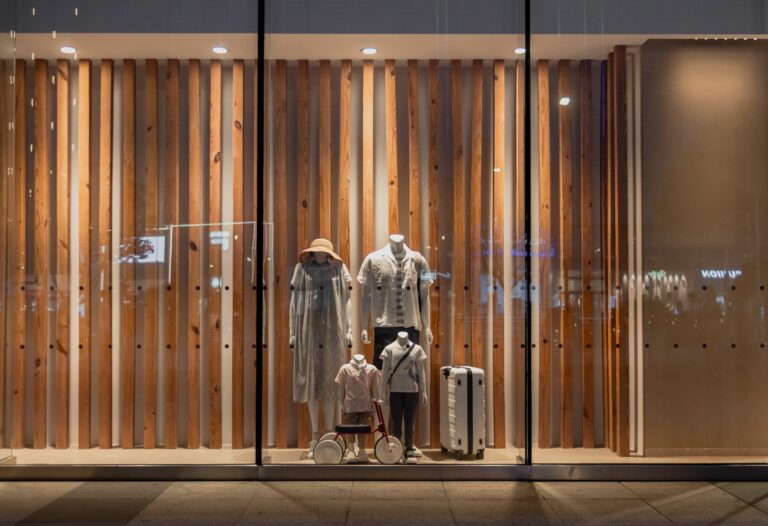 Storefront window display with vertical wooden slats, mannequins in light clothing, and travel luggage behind glass at night.