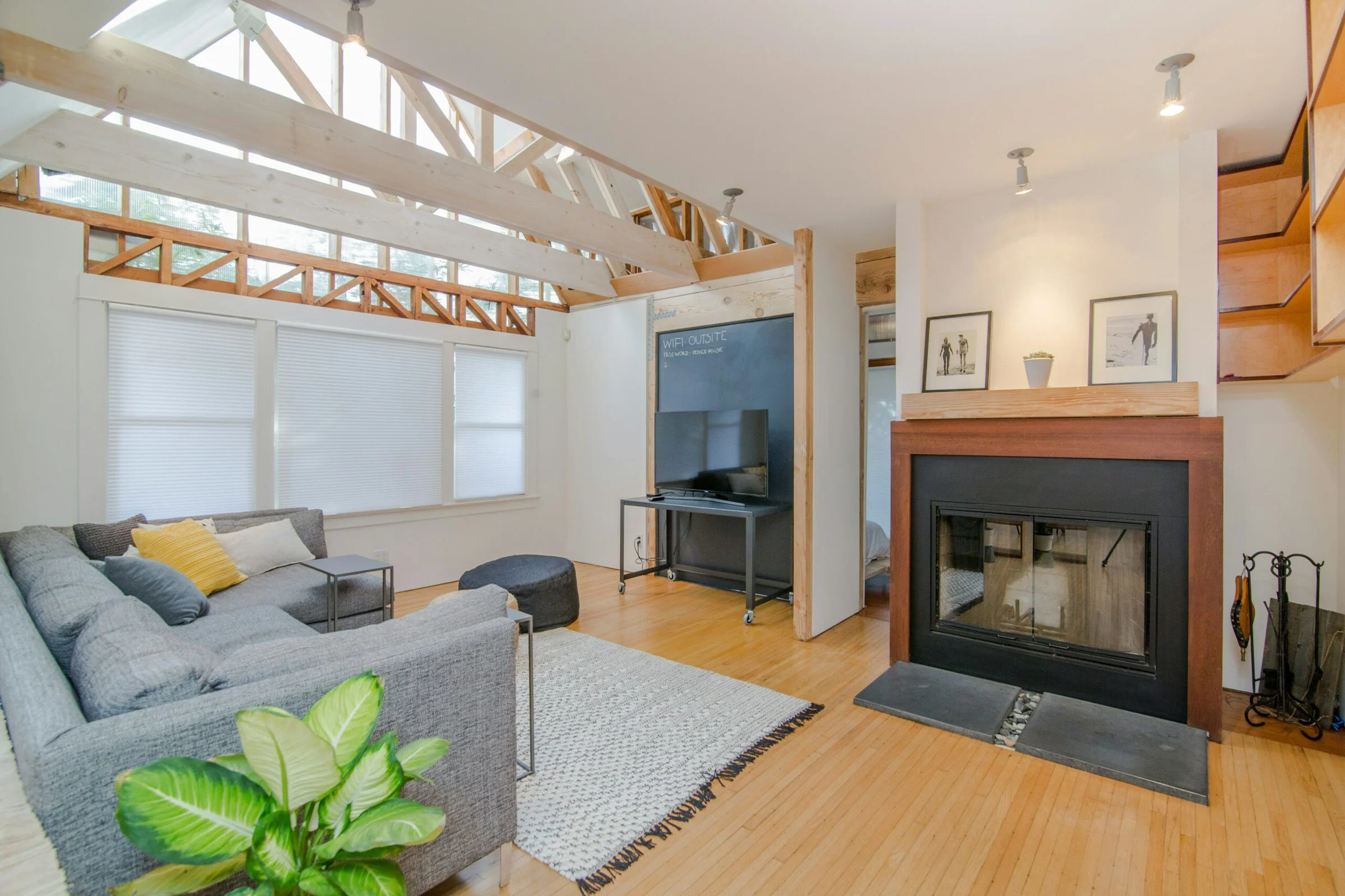 Bright living room with exposed light wood beams, vaulted ceilings, large windows with blinds, and a gray sectional sofa. A fireplace and framed photos sit on the right.